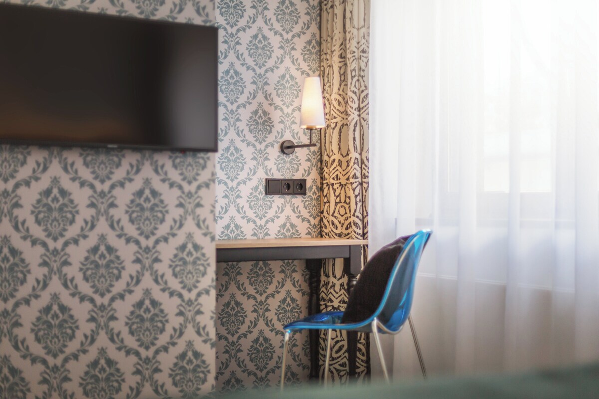 A hotel room with patterned wallpaper, a flat-screen TV mounted on the wall, a desk with a lamp, and a blue chair.
