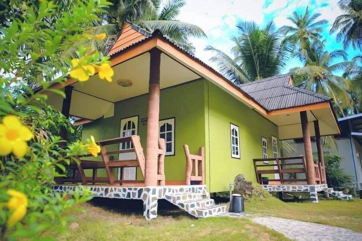 A green house with a tiled porch, wooden furniture, and a stone pathway.