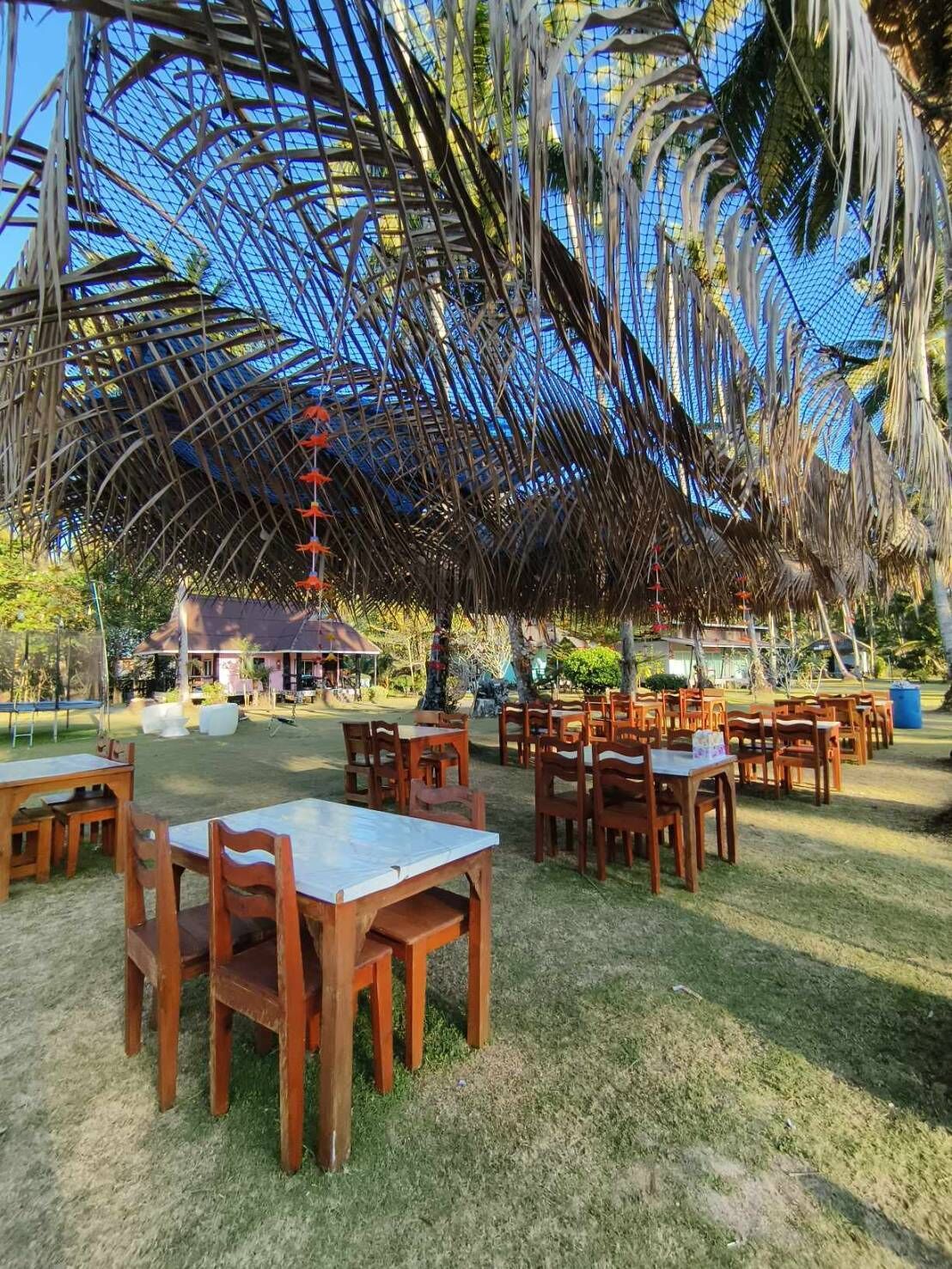 An outdoor dining area with wooden tables and chairs, surrounded by a thatched roof structure and palm trees.