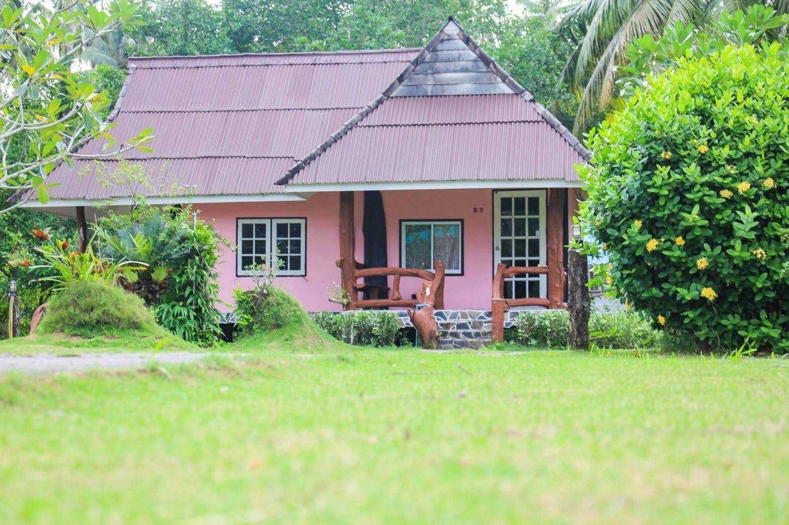 A pink house with a tiled roof, surrounded by lush greenery and a wooden fence.