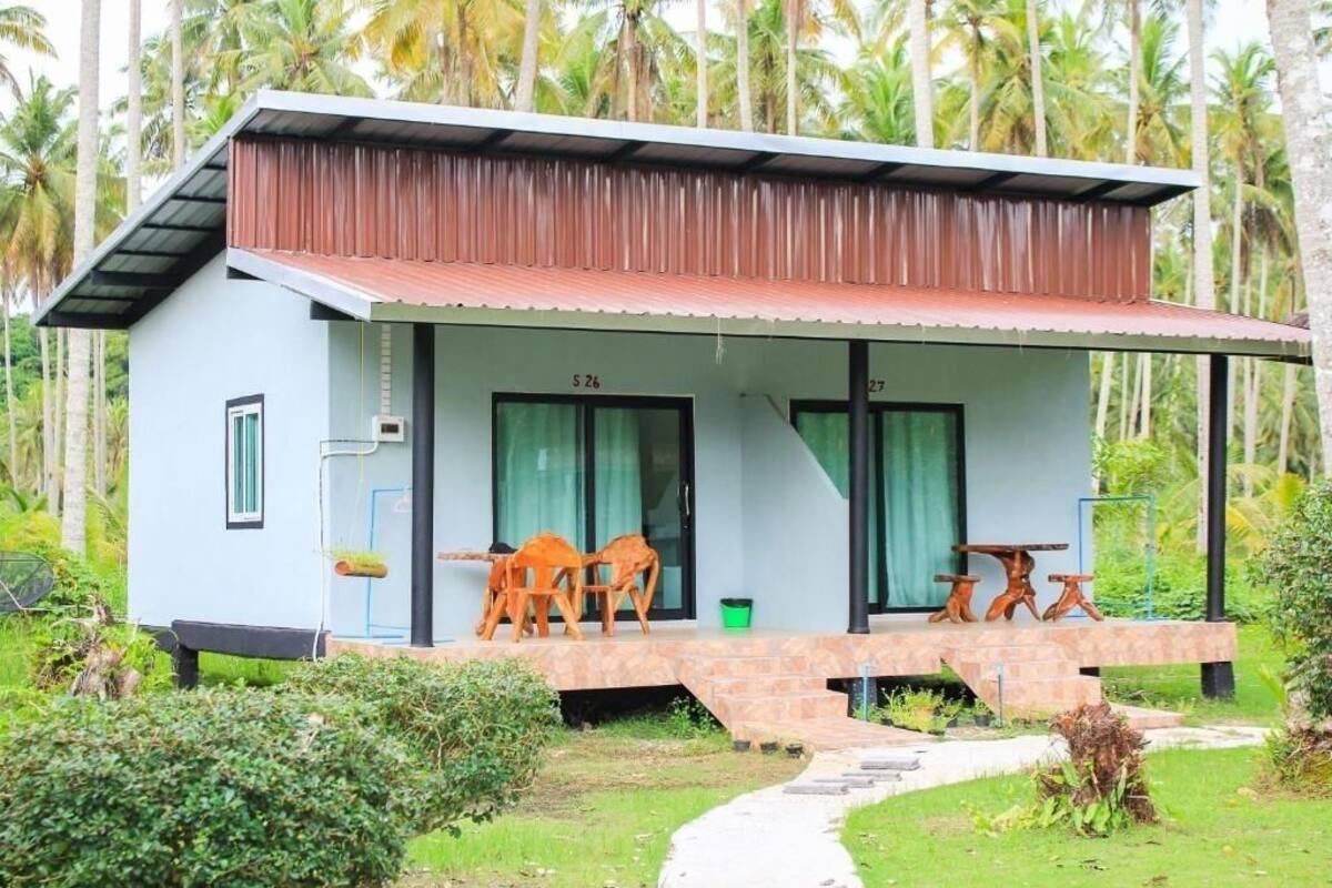 A small, single-story building with a corrugated metal roof, numbered "S 26" and "S 27". It features a porch with wooden chairs and a small table, surrounded by lush greenery and palm trees.