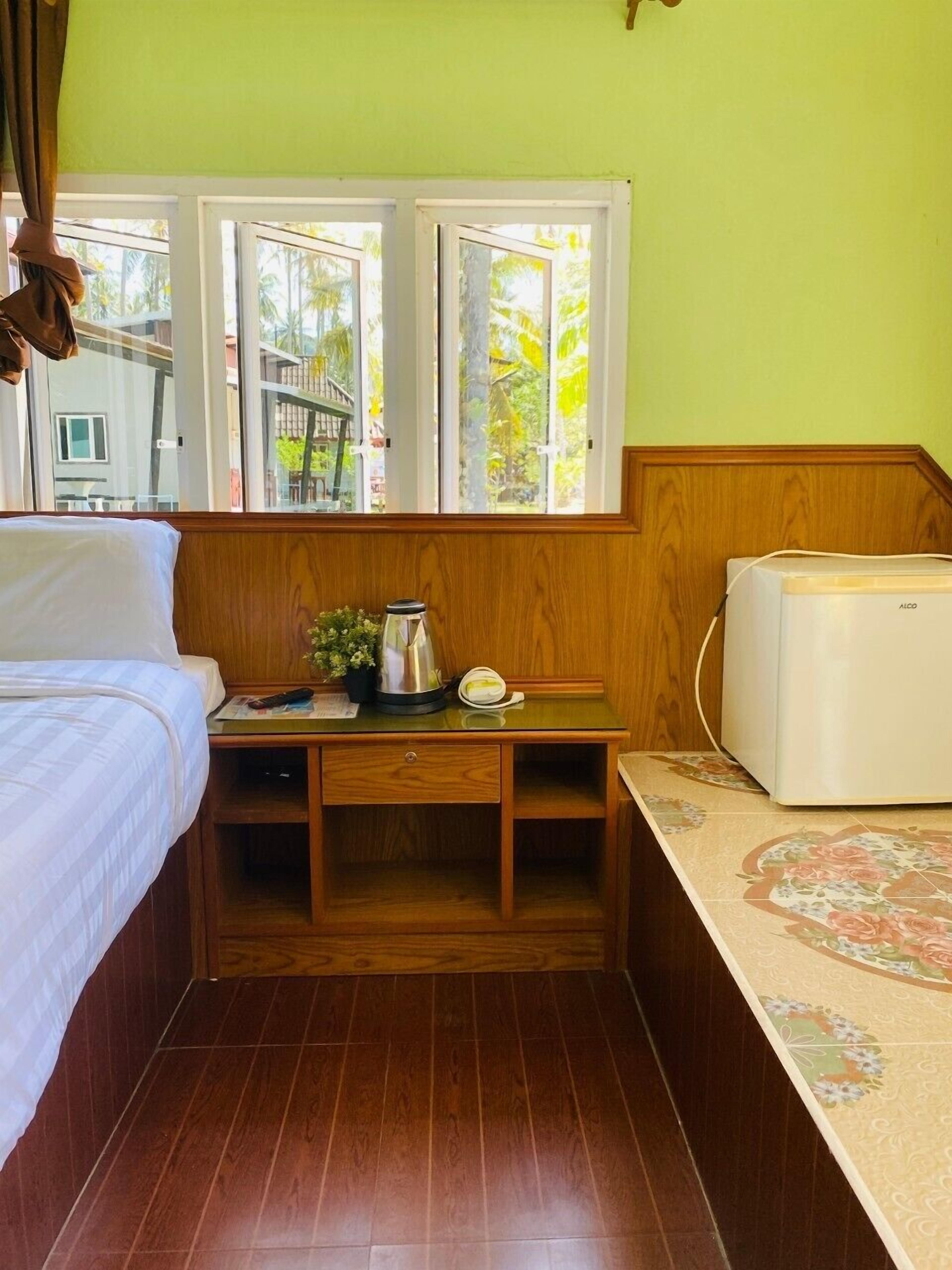 A compact hotel room with a bed, a small wooden table with a kettle and a phone, a microwave, and a window with a view of palm trees.