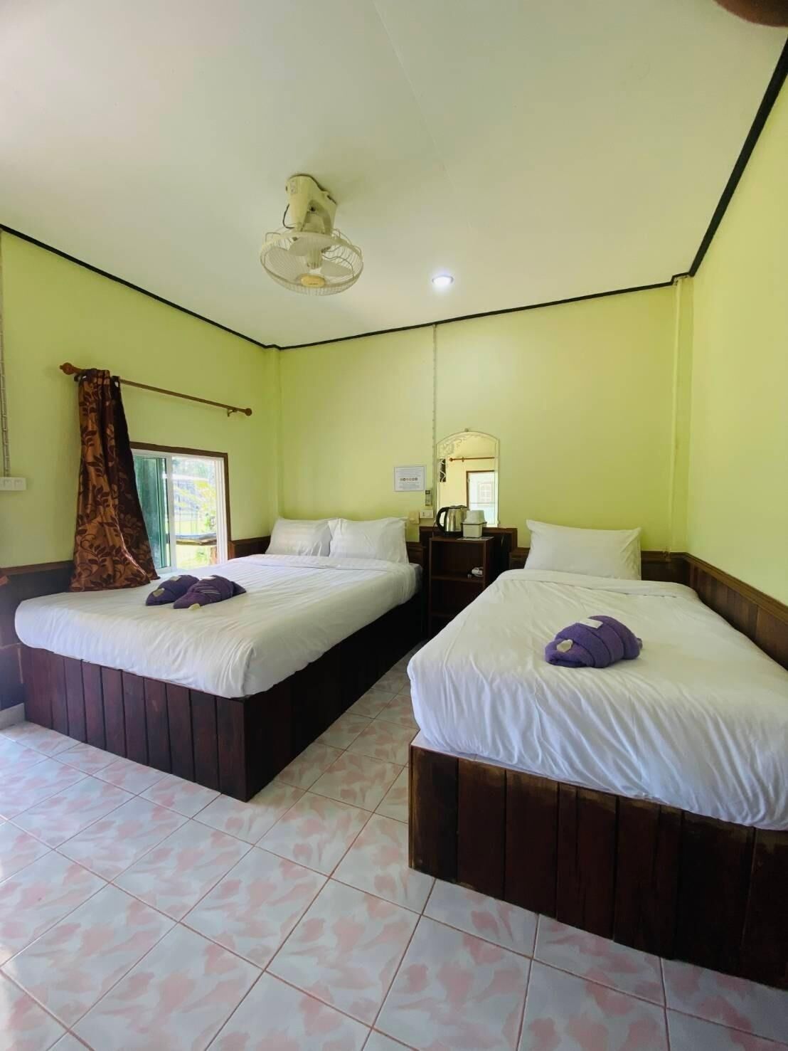Quarto de hotel com duas camas, um ventilador de teto, uma janela com cortinas e uma mesa de cabeceira com abajur.