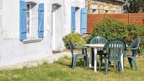 Un bâtiment blanc avec des volets bleus, un mur de pierre et une clôture en bois. On y voit une table entourée de chaises devant le bâtiment.