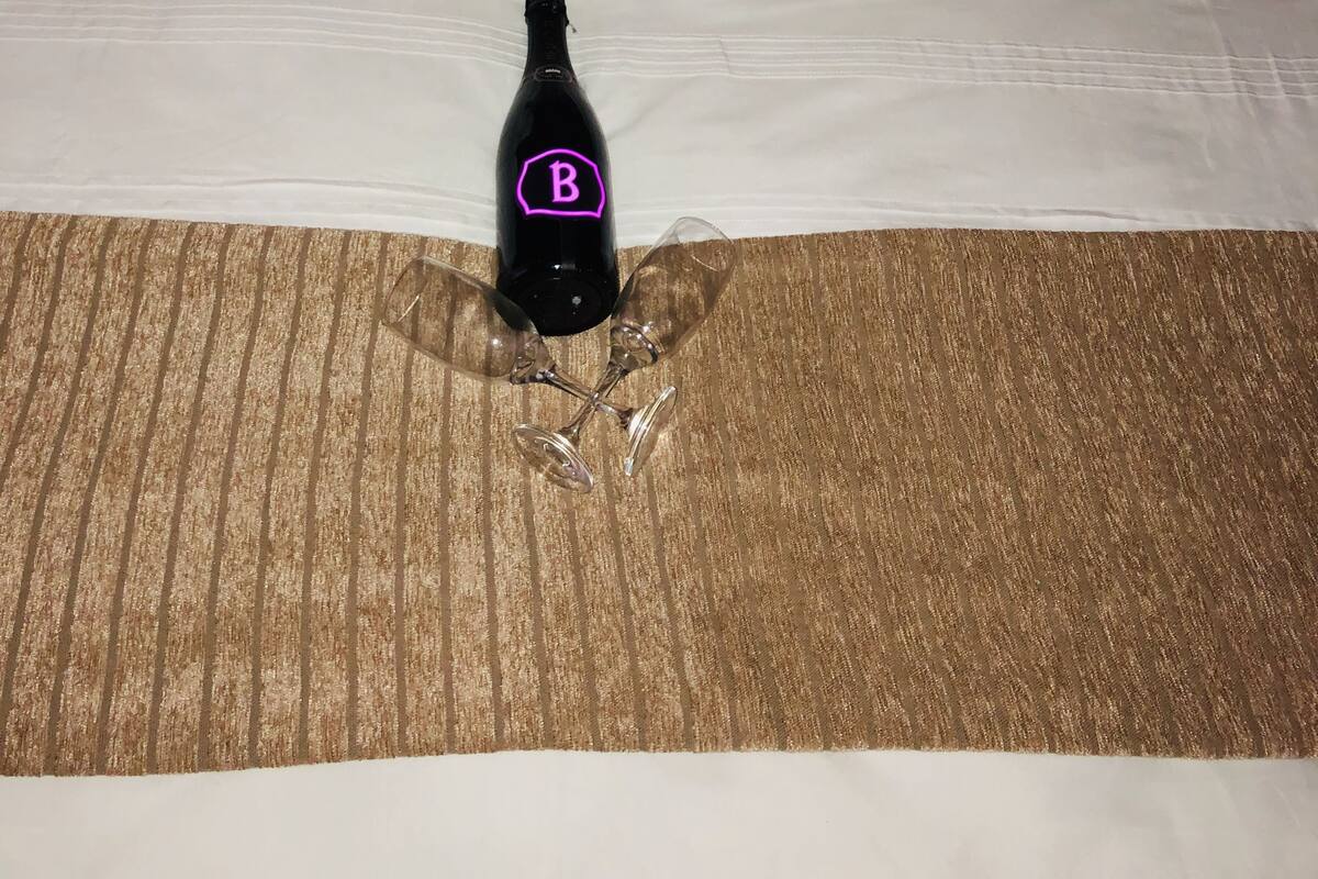 Una cama con un edredón blanco, una almohada decorativa, una toalla blanca enrollada y una botella de champán con un logo 'B' sobre la cama.