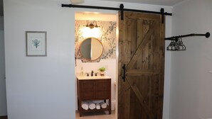 A bedroom with a bed, a wooden door, a bathroom with a sink, mirror, and towels. - Perfect Maine Getaway – Walk to Downtown, Cozy for All - Flexible Cancellation. (Kennebunk)