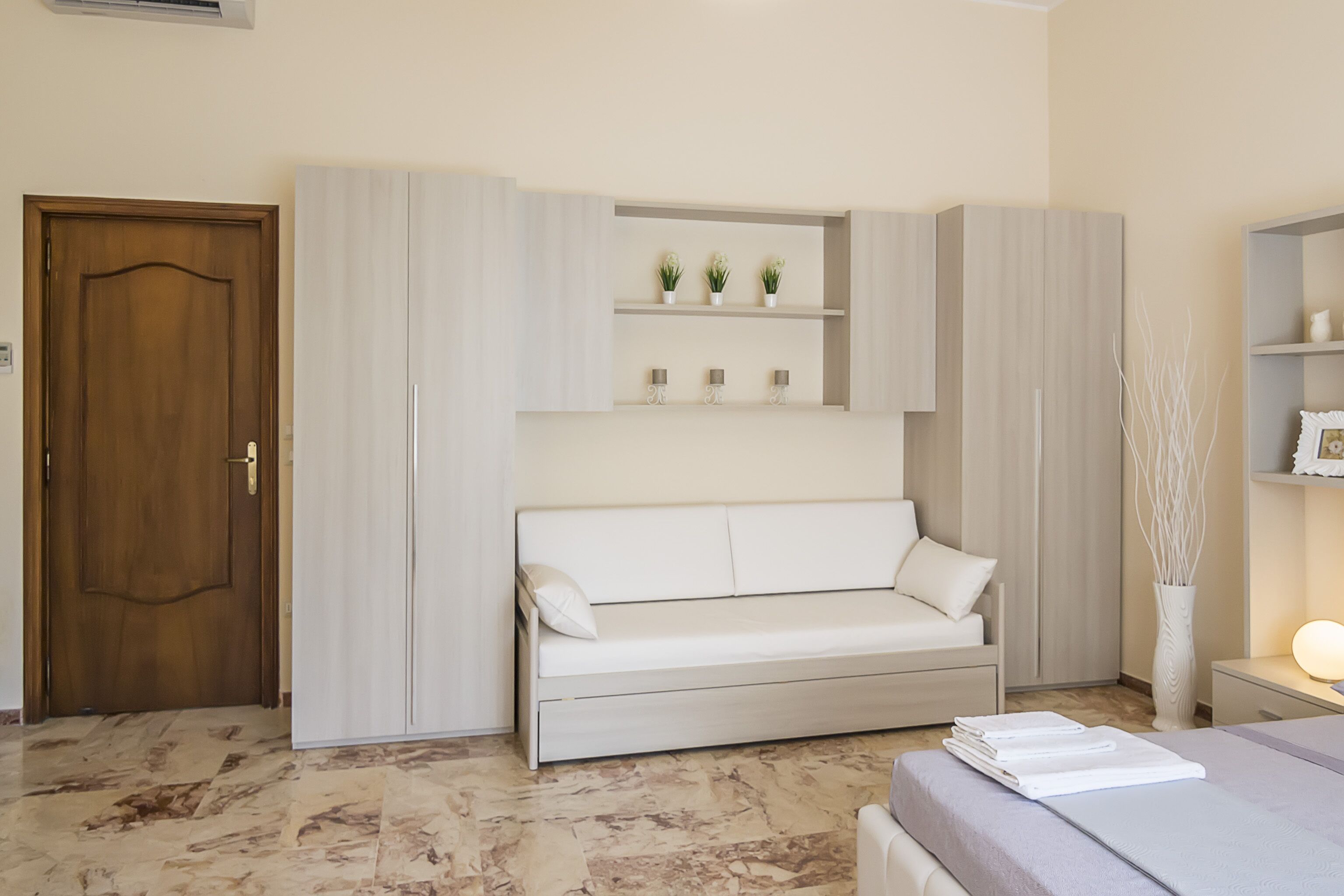 Una camera da letto moderna con un divano letto, mensole a incasso con piante, una porta in legno e un comodino con una lampada.
