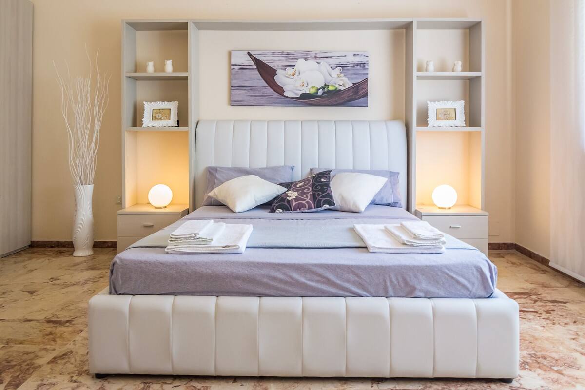 Une chambre à coucher bien rangée, avec un grand lit, deux tables de chevet avec des lampes, un vase décoratif et un tableau encadré au mur.