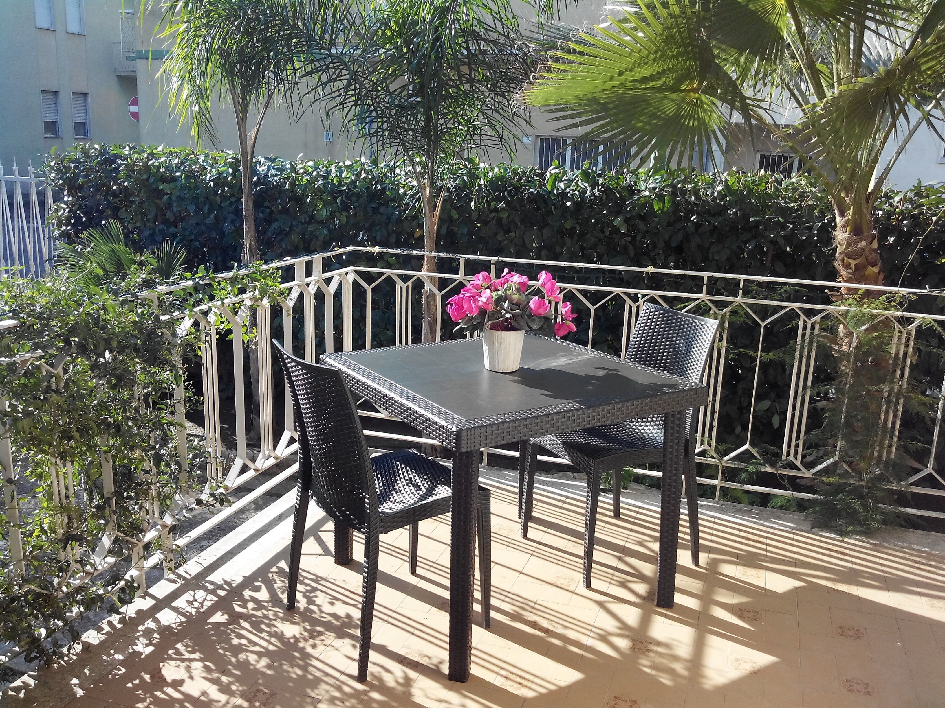An outdoor patio with a table and chairs, surrounded by a white metal fence, and various plants and trees in the background.