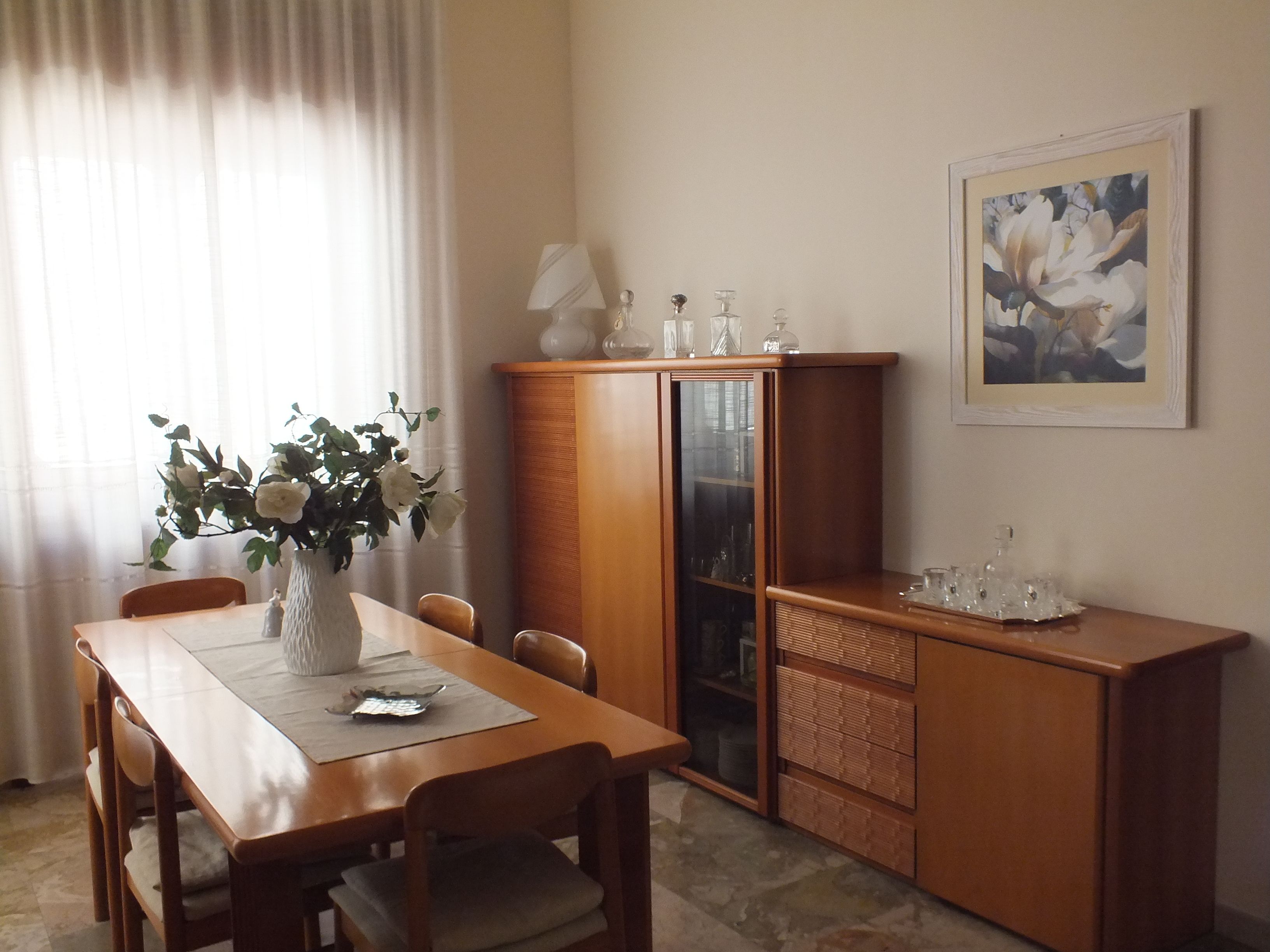 A dining area with a wooden table and chairs, a vase with flowers, a cabinet with glass doors, and a framed painting on the wall.