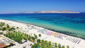 A beach with rows of umbrellas and sun loungers, a parking area, and a clear view of the sea and distant mountains.