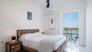 Quarto com cama de madeira, roupa de cama branca, luminária de cabeceira, ar condicionado e varanda com mobiliário de exterior.