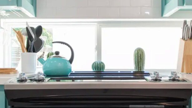 Una cocina con una campana extractora de acero inoxidable, una ventana con vista a un cactus y un hervidor azul verdoso sobre la encimera.