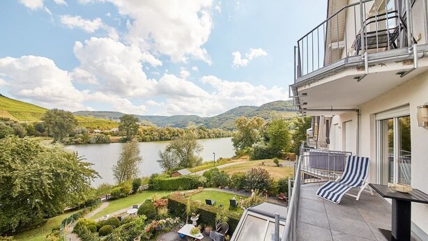 Un balcón con vistas a un lago y colinas, un pequeño jardín y una mesa de comedor con sillas.