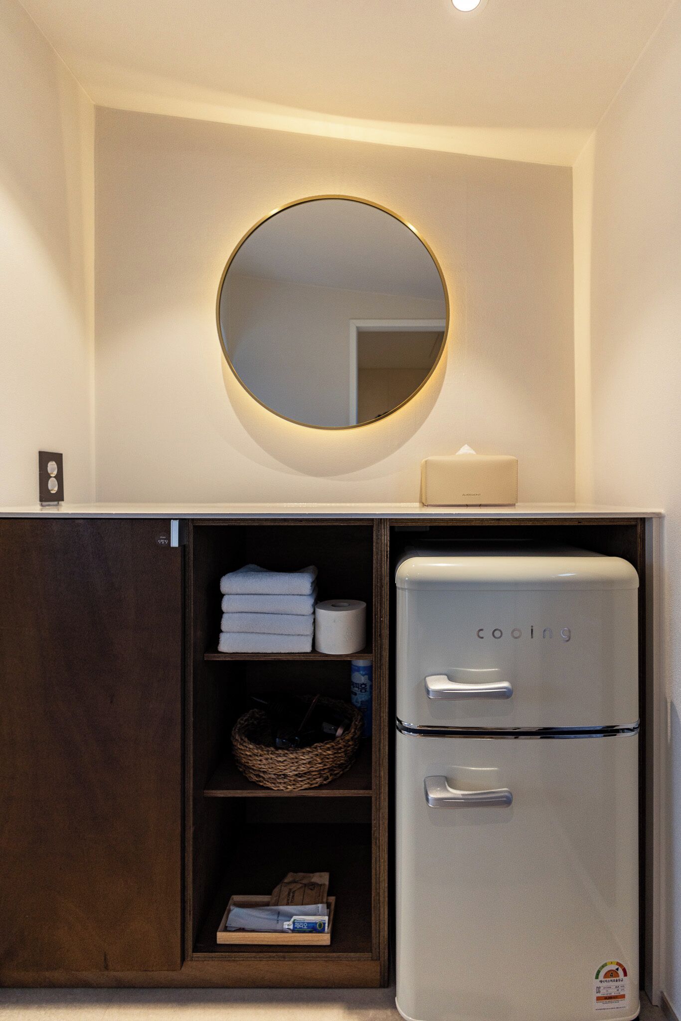 A compact kitchenette with a white refrigerator, a built-in oven, a mirror, and a shelf with towels and a basket.