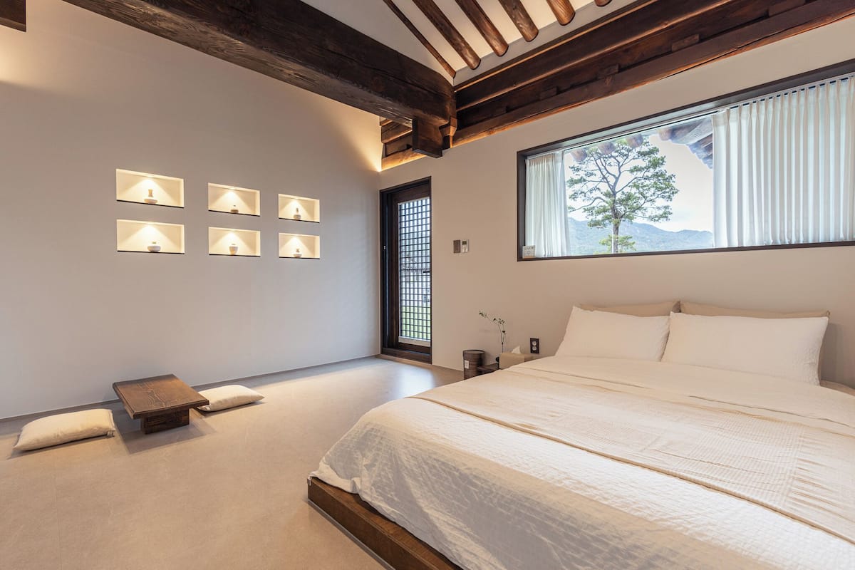 A bedroom with a large bed, a small table, a window with a view of trees, and wall-mounted lights.