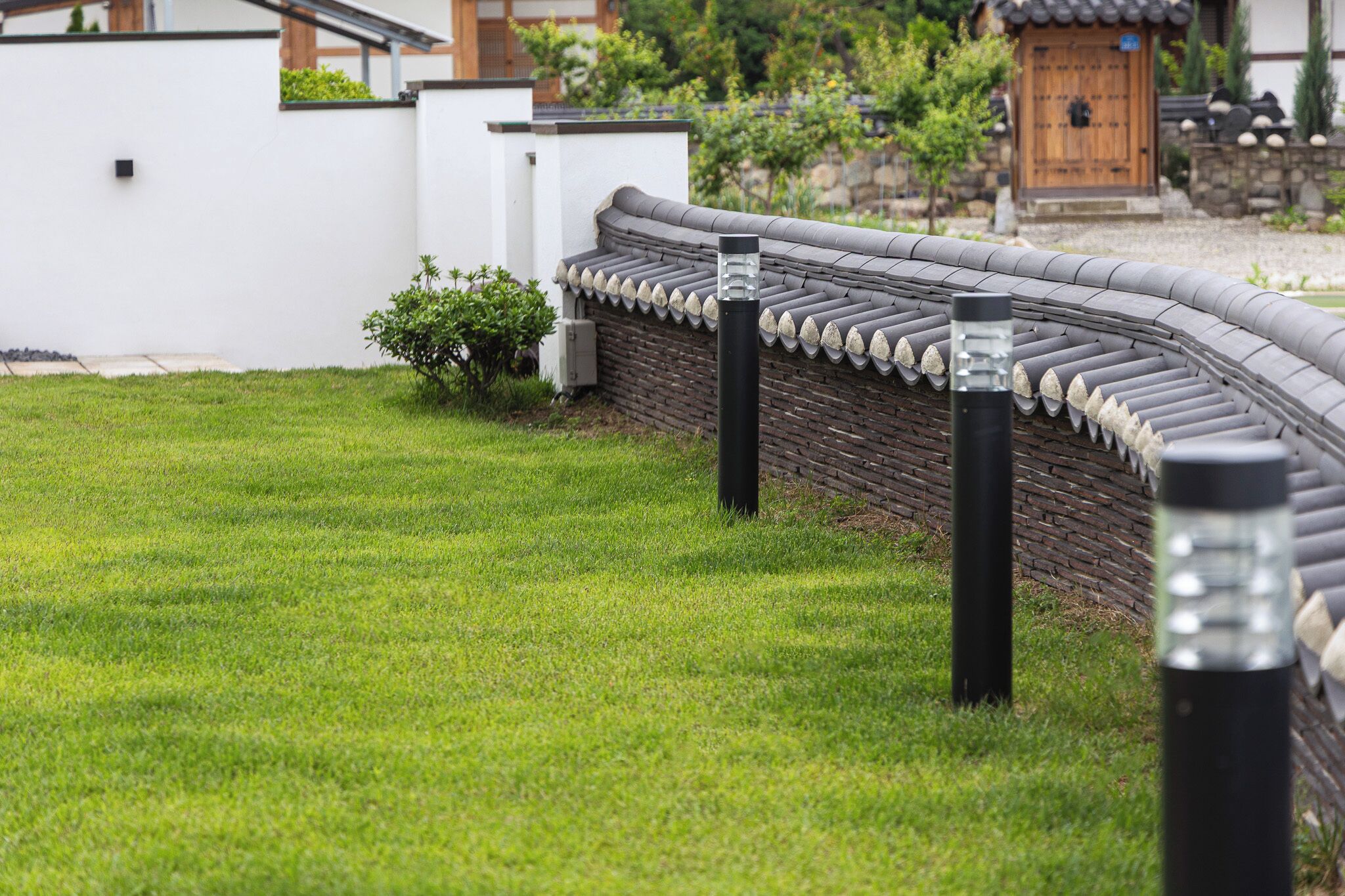 Un bâtiment de style japonais traditionnel, doté d’un toit incurvé, d’un mur blanc et d’un petit jardin avec de l’herbe et quelques arbustes.