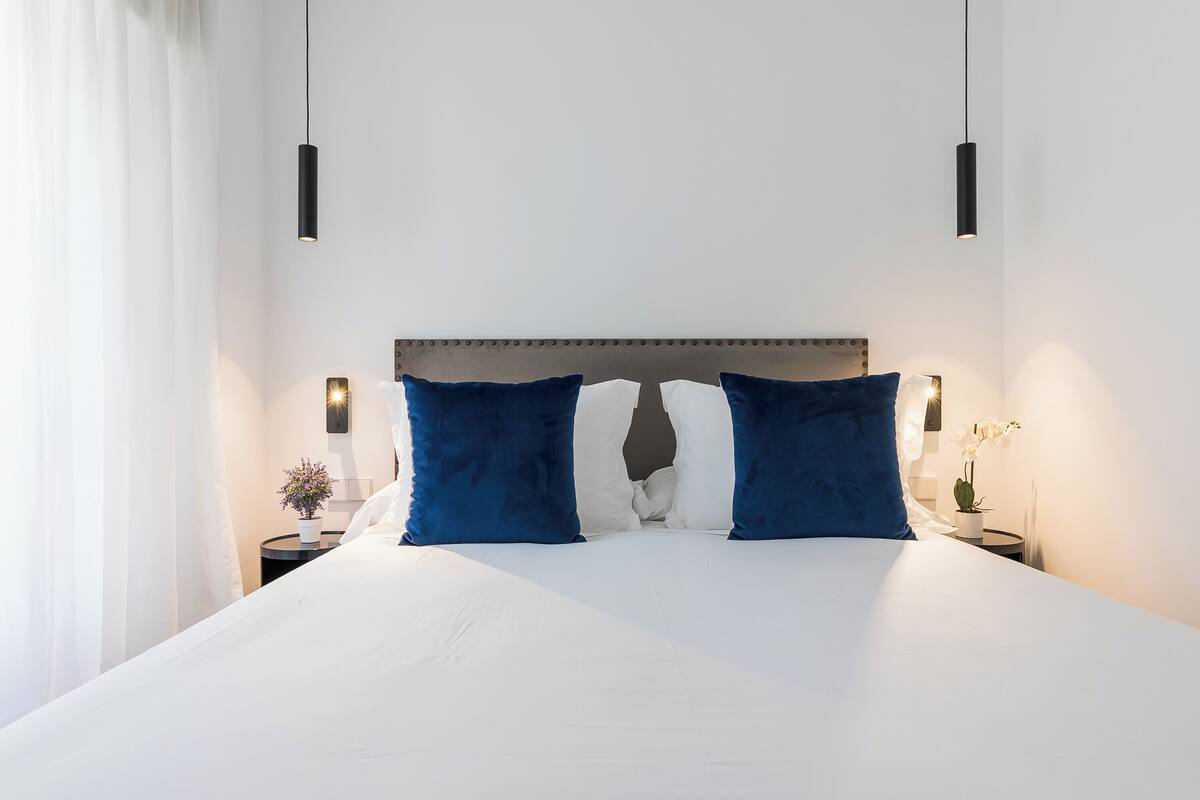 Ein ordentlich bezogenes Bett mit weißen Leinen und zwei blauen Kissen, ein Nachttisch mit einer kleinen Pflanze und eine Wandleuchte.
