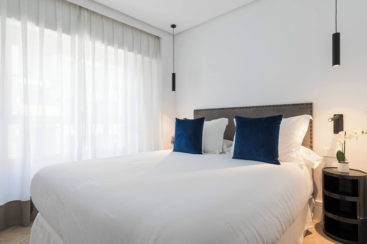 Ein ordentlich bezogenes Bett mit weißer und blauer Bettwäsche, ein Nachttisch mit Lampe sowie ein Fenster mit leichtem Vorhang.