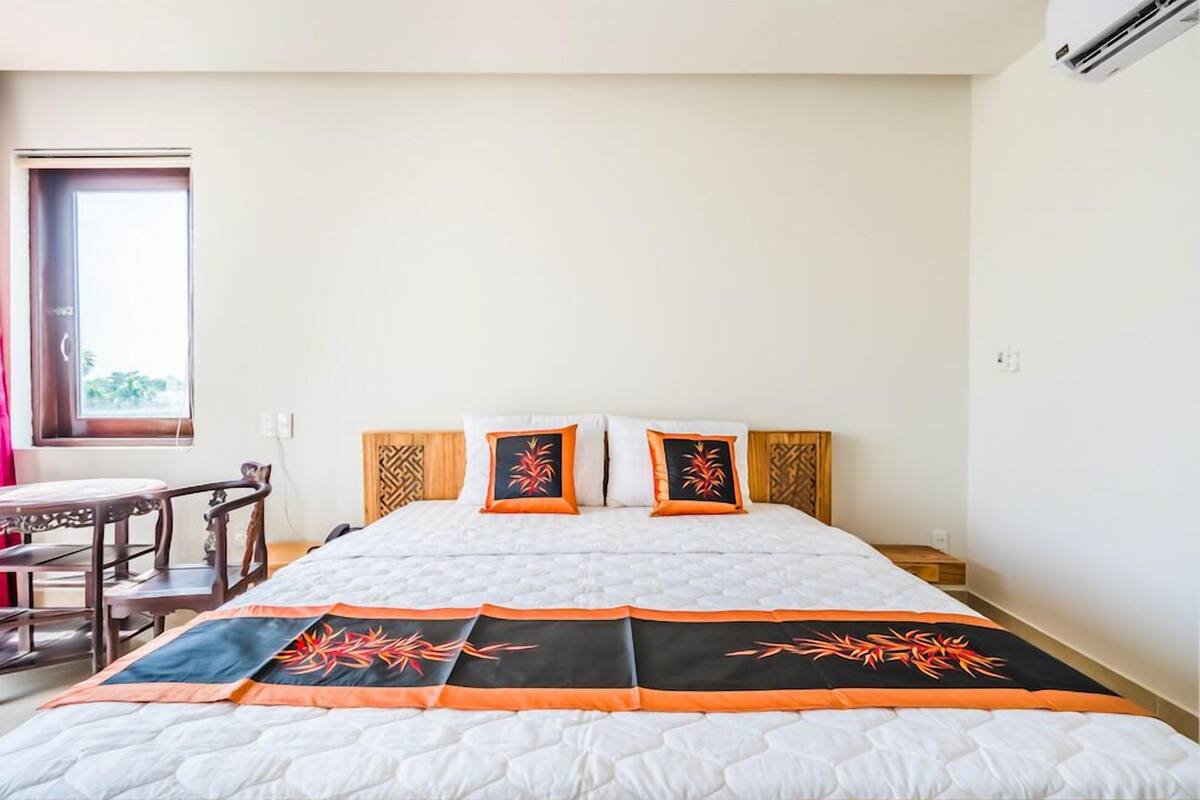 A hotel room with a bed, wooden headboard, two pillows, a bedside table, and a window with a view.