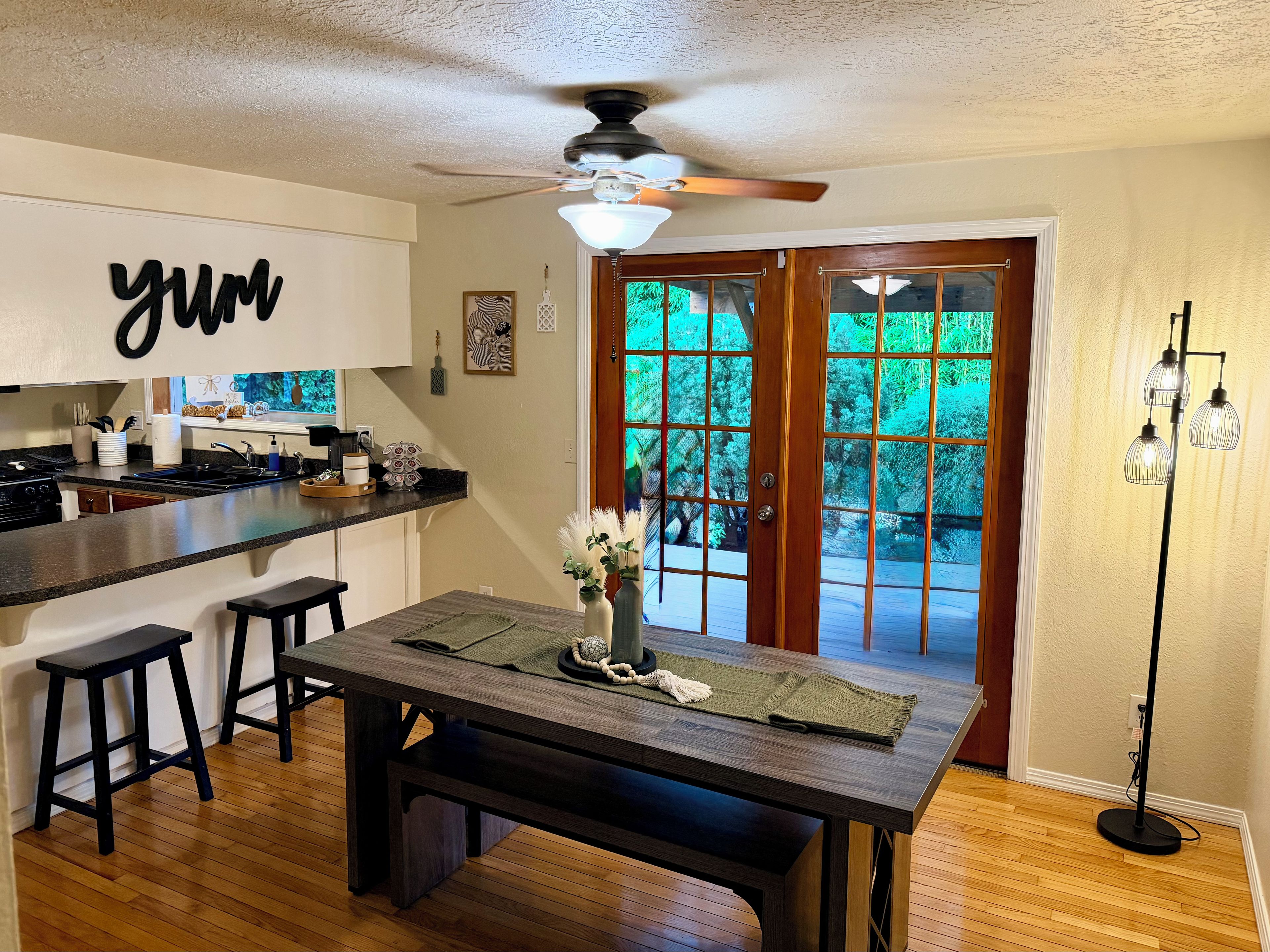 A kitchen with a bar area, wooden flooring, a ceiling fan, and a view through a glass door.