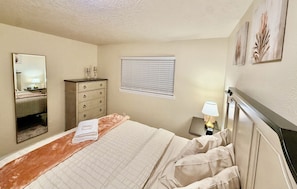 A hotel room with a bed, a dresser, a mirror, a window with blinds, and a nightstand with a lamp.