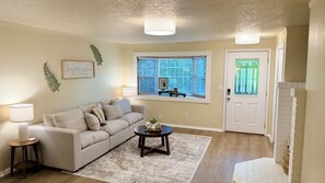A living room with a sofa, a coffee table, a lamp, a window with blinds, and a door.