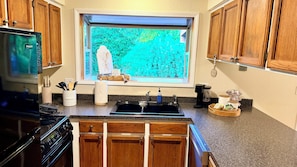 A kitchen with wooden cabinets, a stainless steel refrigerator, a window with a view of greenery, a sink, and various kitchen items.