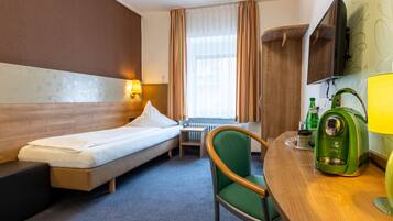 A hotel room with a bed, a chair, a small table, a coffee machine, and a TV mounted on the wall.