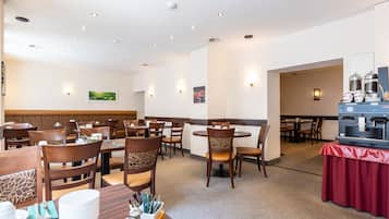 A hotel dining area with multiple tables and chairs, a coffee station, and wall-mounted light fixtures.