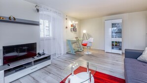 A modern living room with a flat-screen TV, a glass coffee table, a red rug, a white sofa, a dining table with a chair, and a bookshelf. - Fiorello Apartment (Rijeka)