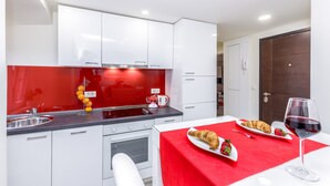 A modern kitchen with white cabinets, a red backsplash, and a dining area with a red tablecloth. There is a croissant, a strawberry, and a glass of red wine on the table. - Fiorello Apartment (Rijeka)
