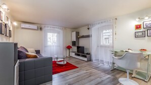 A modern living room with a grey sofa, a glass coffee table, and a red rug. There is a TV, a bookshelf, and a dining area with a glass table and white chair. - Fiorello Apartment (Rijeka)