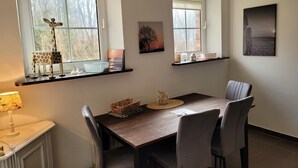 A dining area with a wooden table and four chairs, two windows with curtains, a lamp, and a shelf with decorative items. - Apartment "Dat Nüst", terrace, garden, mod. bath (Manslagt)