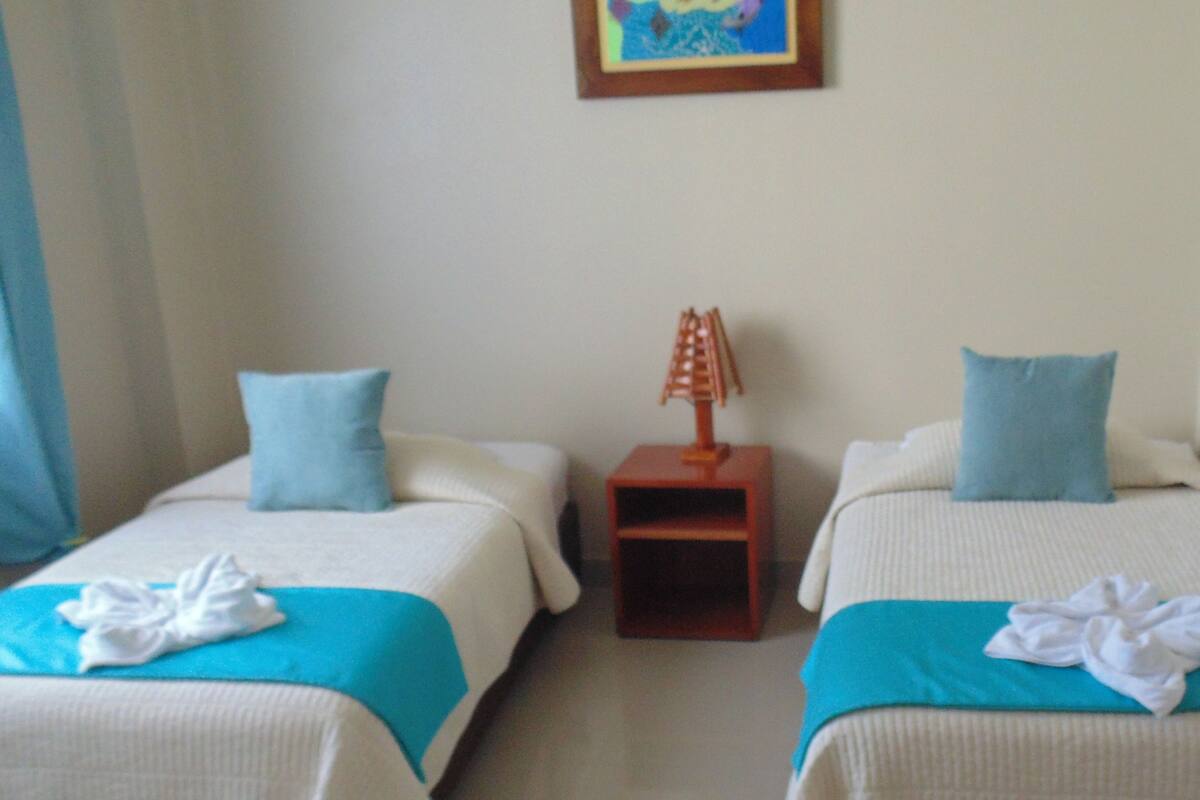 Two single beds with blue bedding and white pillows, a small wooden nightstand with a lamp, and a framed picture on the wall.