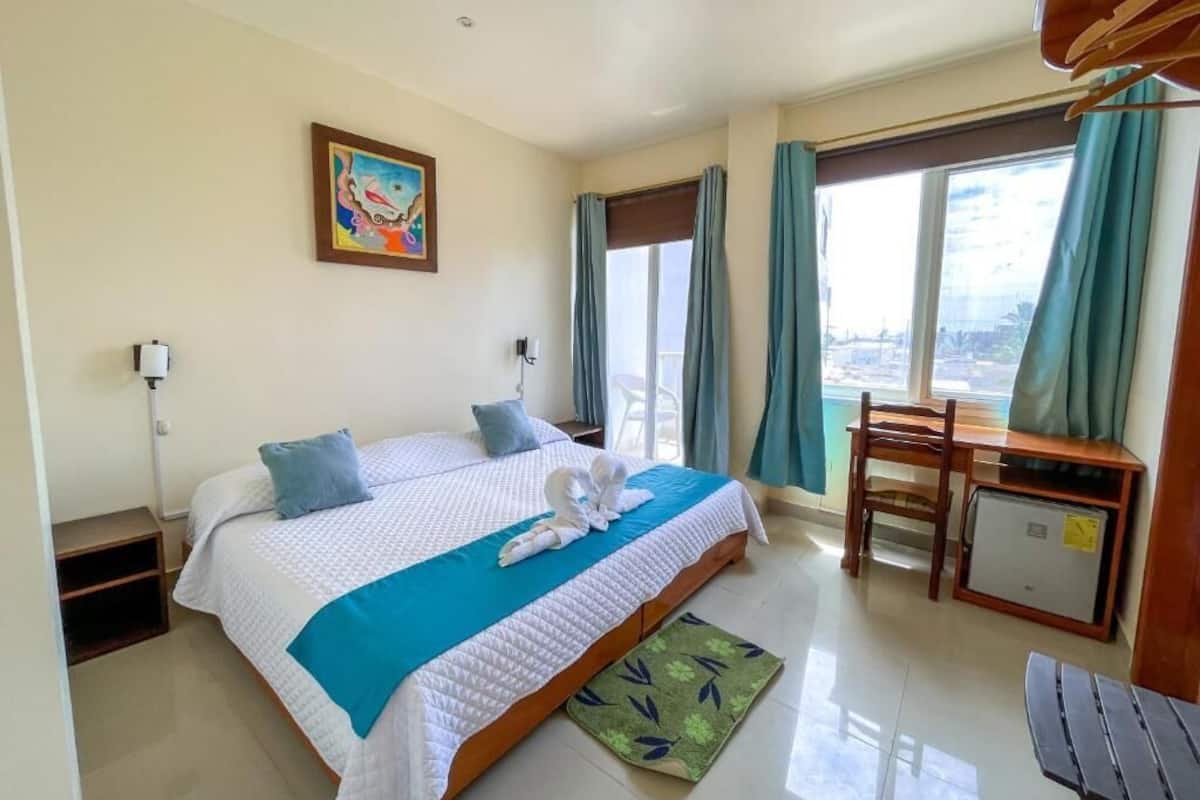 A hotel room with a bed, a desk, a chair, a small table, a window with curtains, and a painting on the wall.