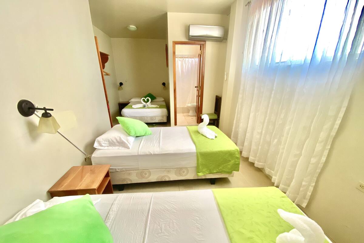 A hotel room with two beds, each with a green pillow and a white towel. There is a bedside table with a lamp, a window with white curtains, and an air conditioning unit on the wall.