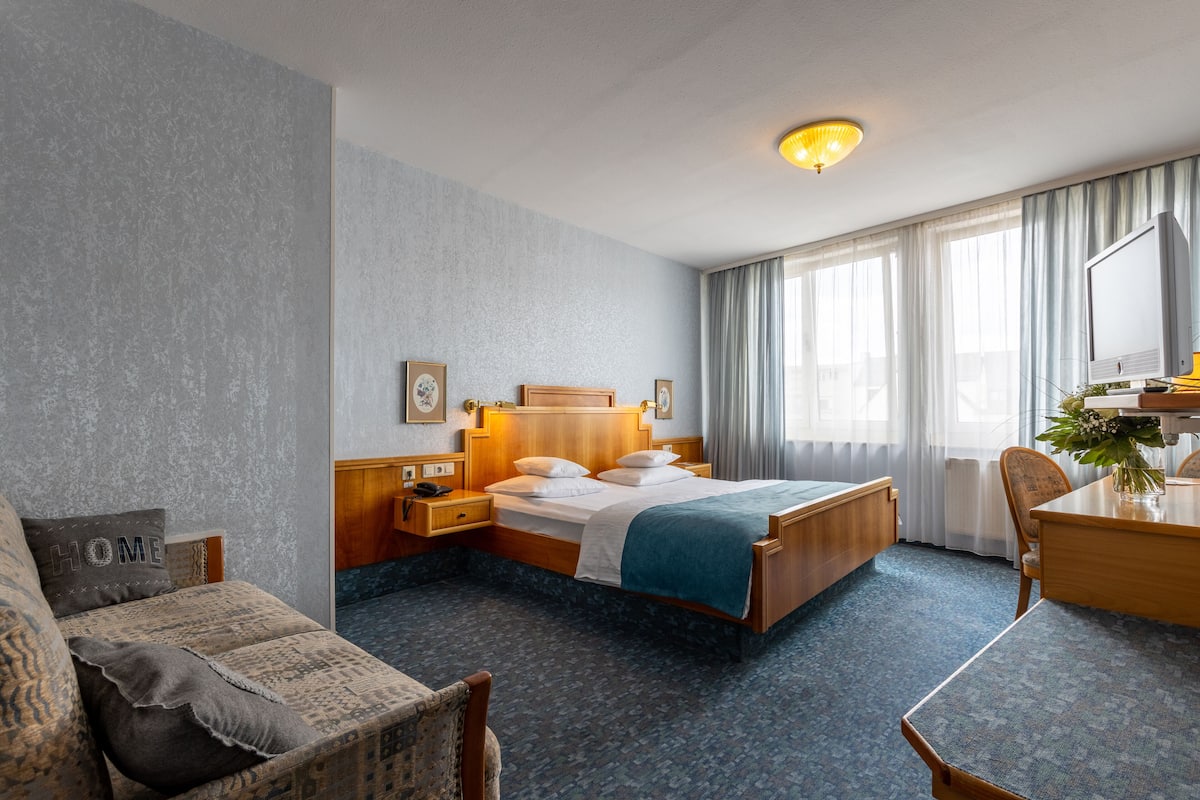 A hotel room with a bed, a desk, a chair, a small table, a lamp, and a window with curtains.