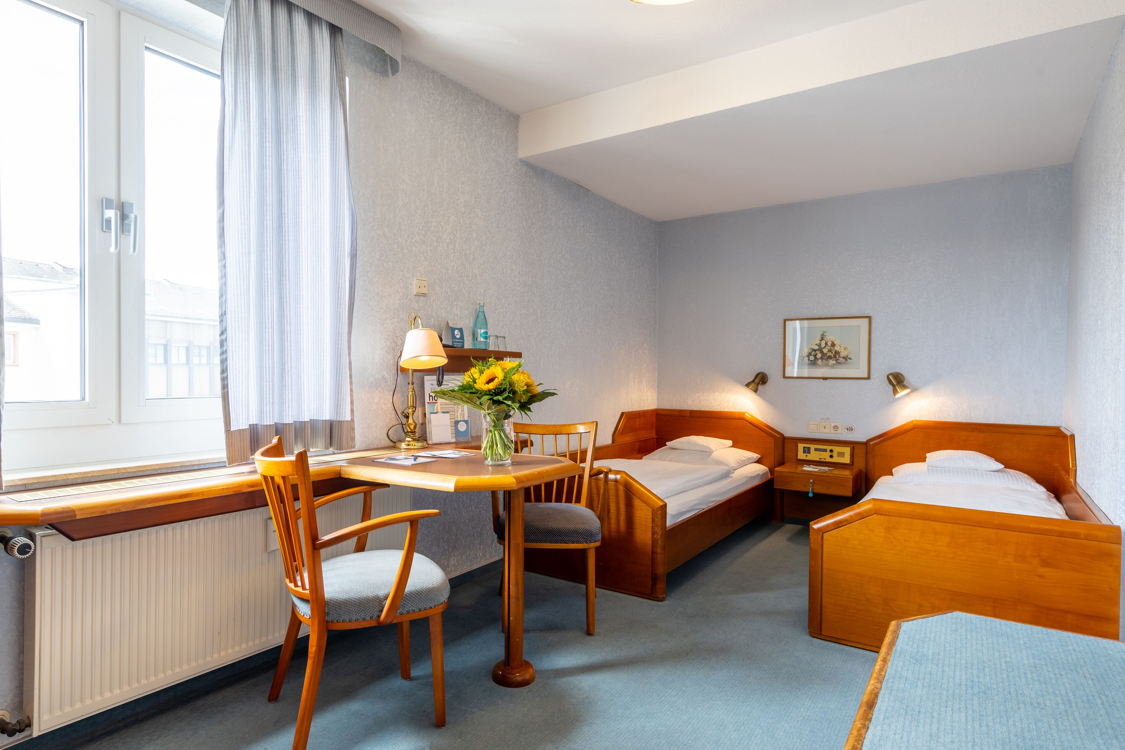 Habitación de hotel con una cama de madera, un escritorio con una silla, una mesita con un jarrón de flores y una ventana con cortinas.
