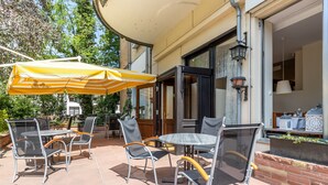 An outdoor patio with metal chairs and a table, a yellow striped umbrella, and a brick wall. - TRIP INN Hotel Aschaffenburger Hof (Aschaffenburg)