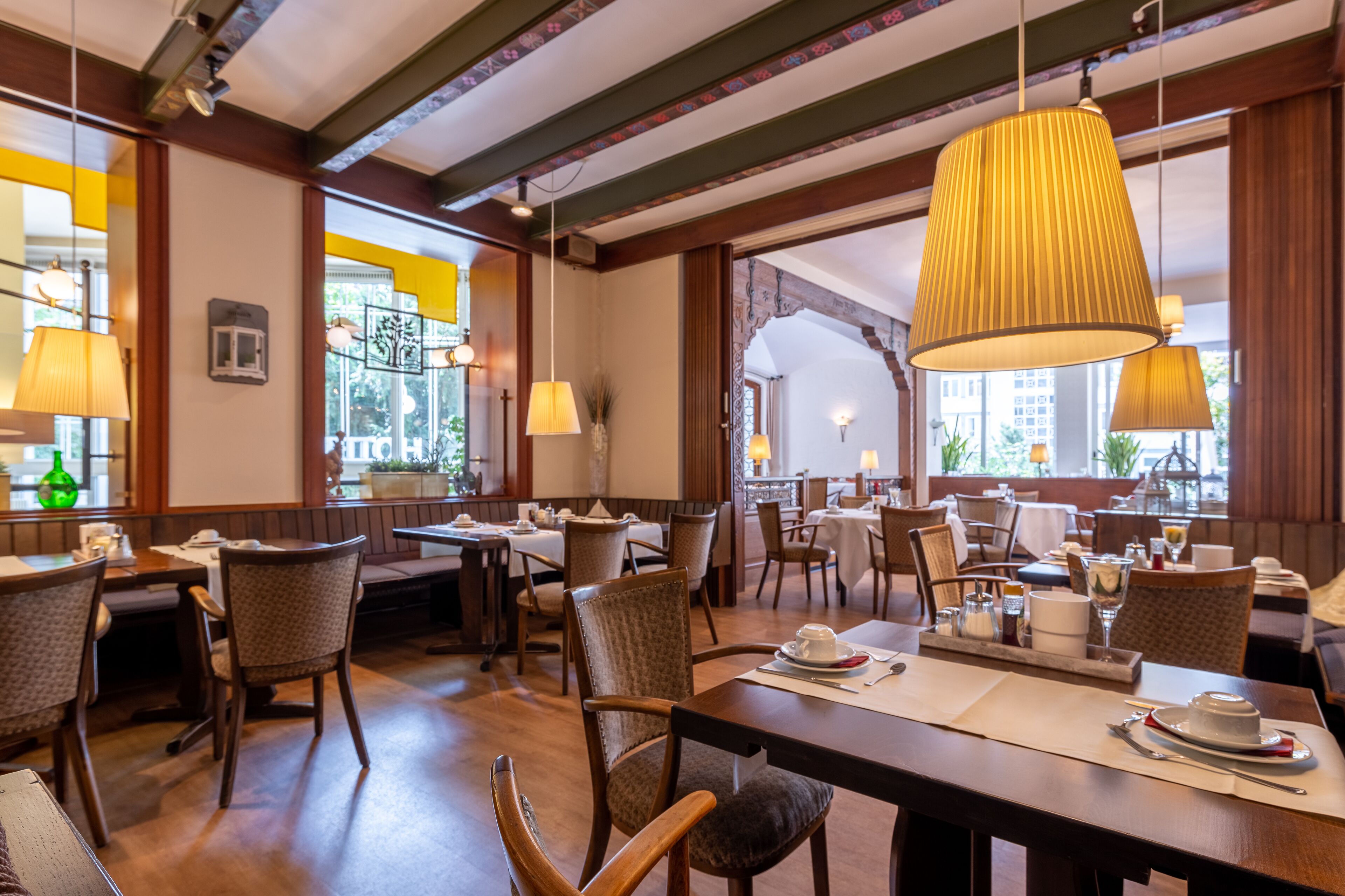 Ein Restaurant mit Holztischen und -stühlen, großen Pendelleuchten und Fenstern mit Blick auf Gebäude.