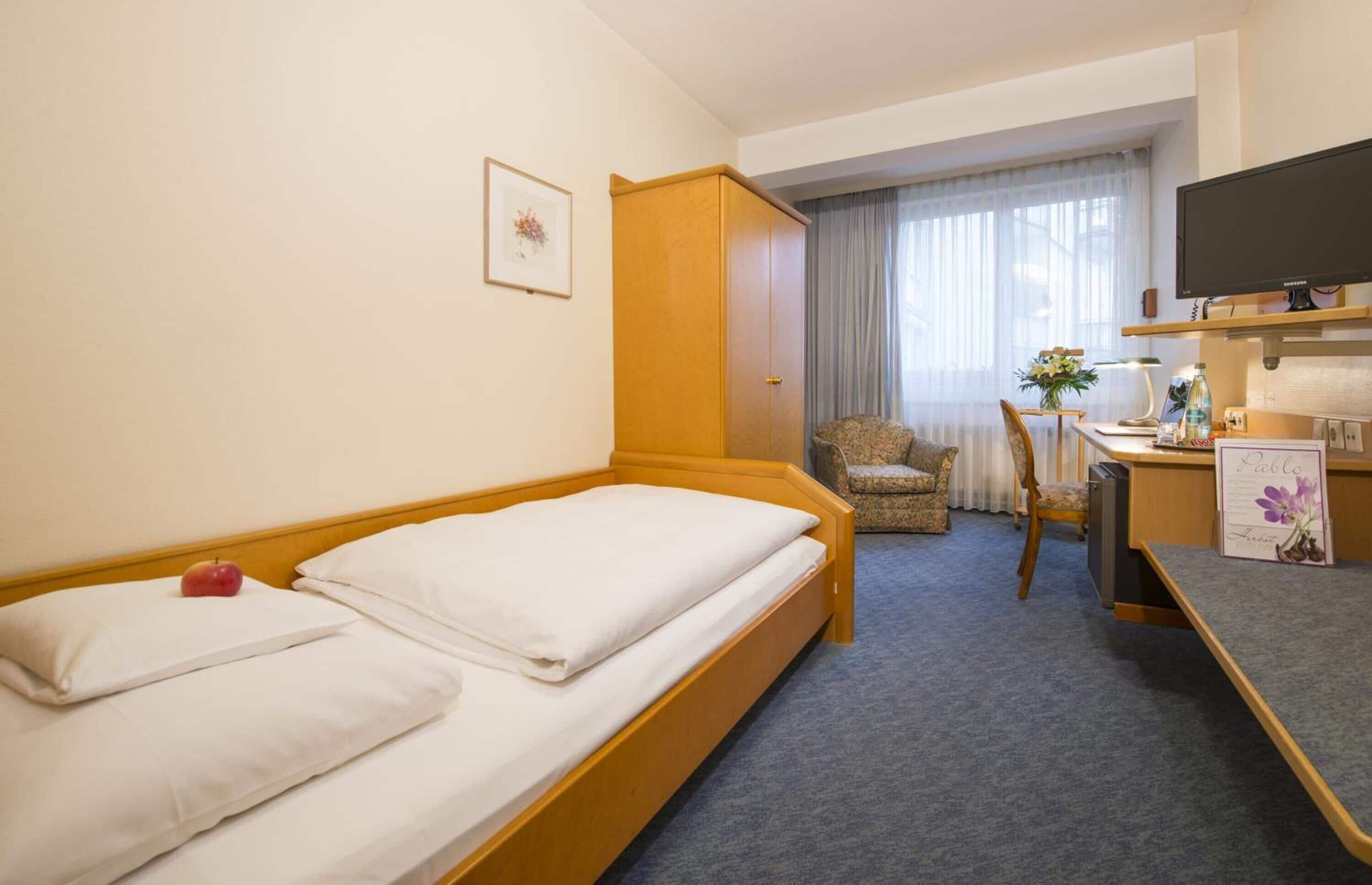 A hotel room with a single bed, a desk with a chair, a television, and a small table with a flower vase.