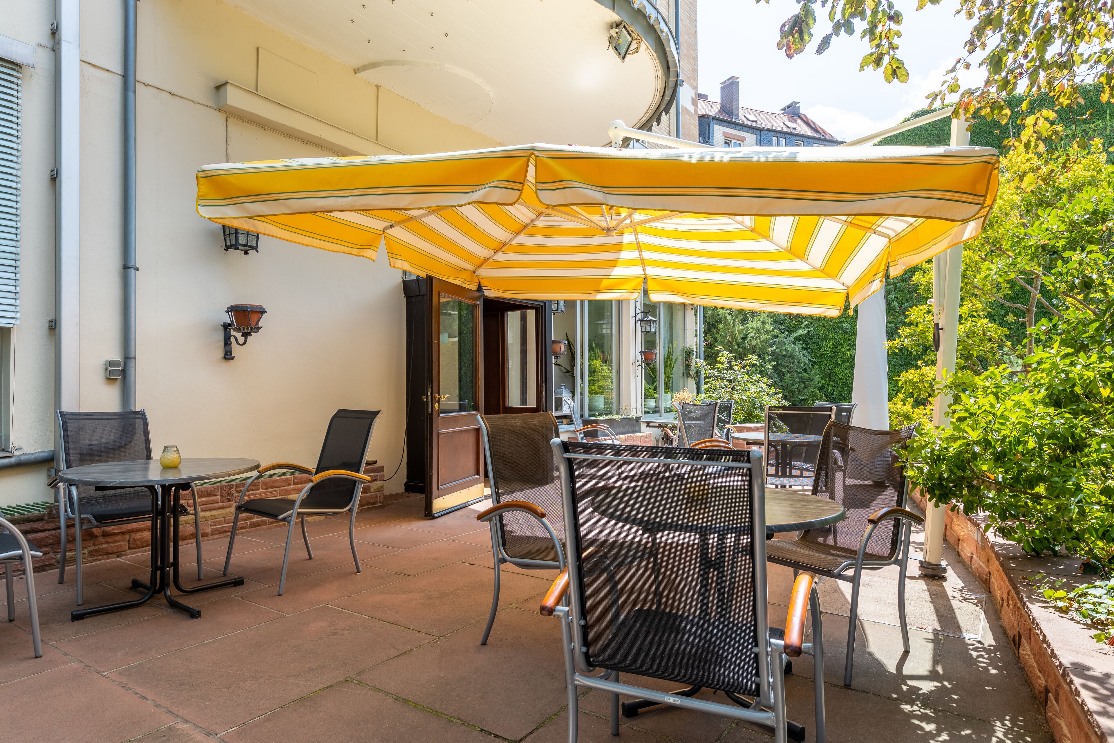 Eine Terrasse im Freien mit Tisch und Stühlen, einem gelb gestreiften Sonnenschirm und Blick auf Grünflächen.
