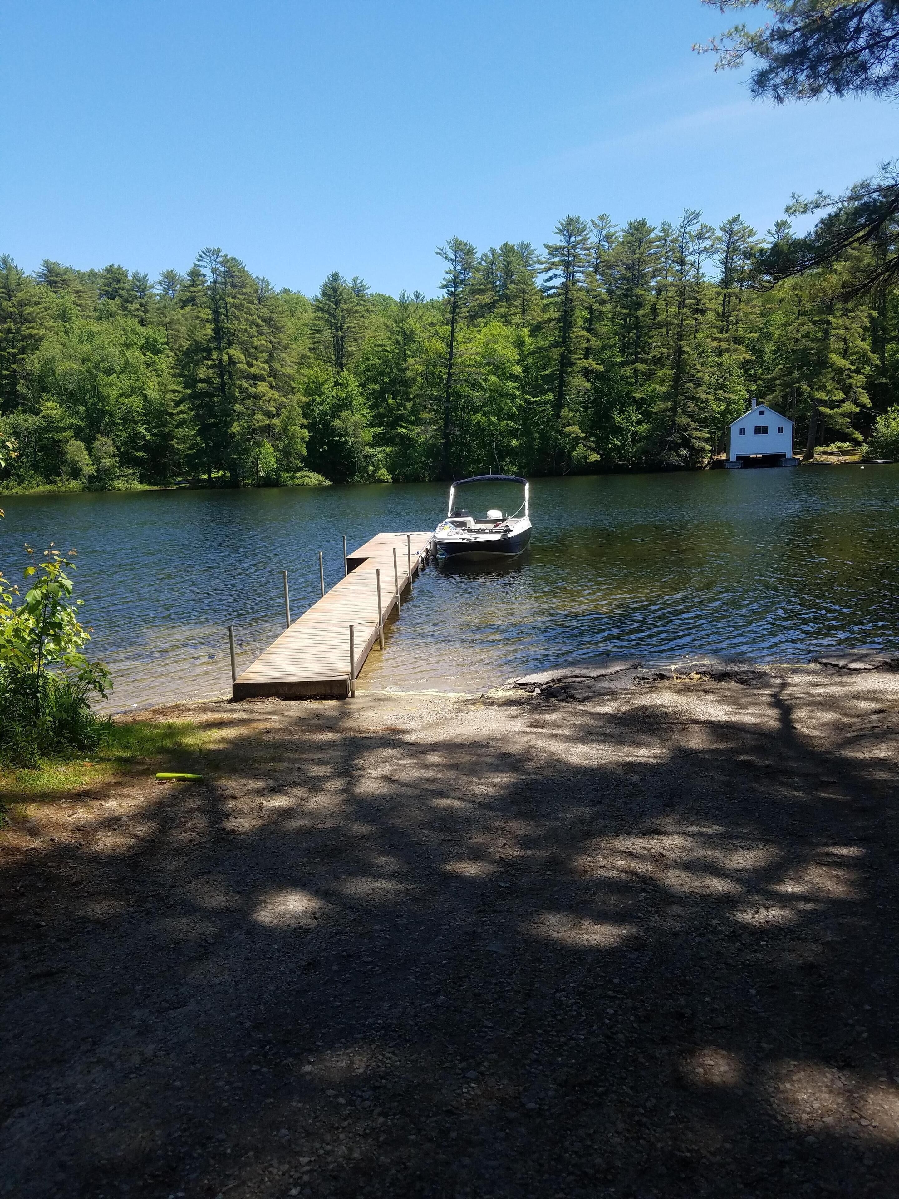 A dock extending into a lake with a boat moored at the end. A small white cabin is visible on the opposite shore.