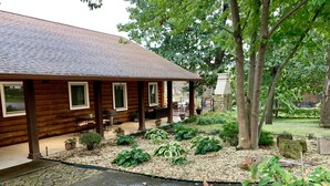 A log cabin with a covered patio, stone pathway, and a garden with various plants and a small pond.