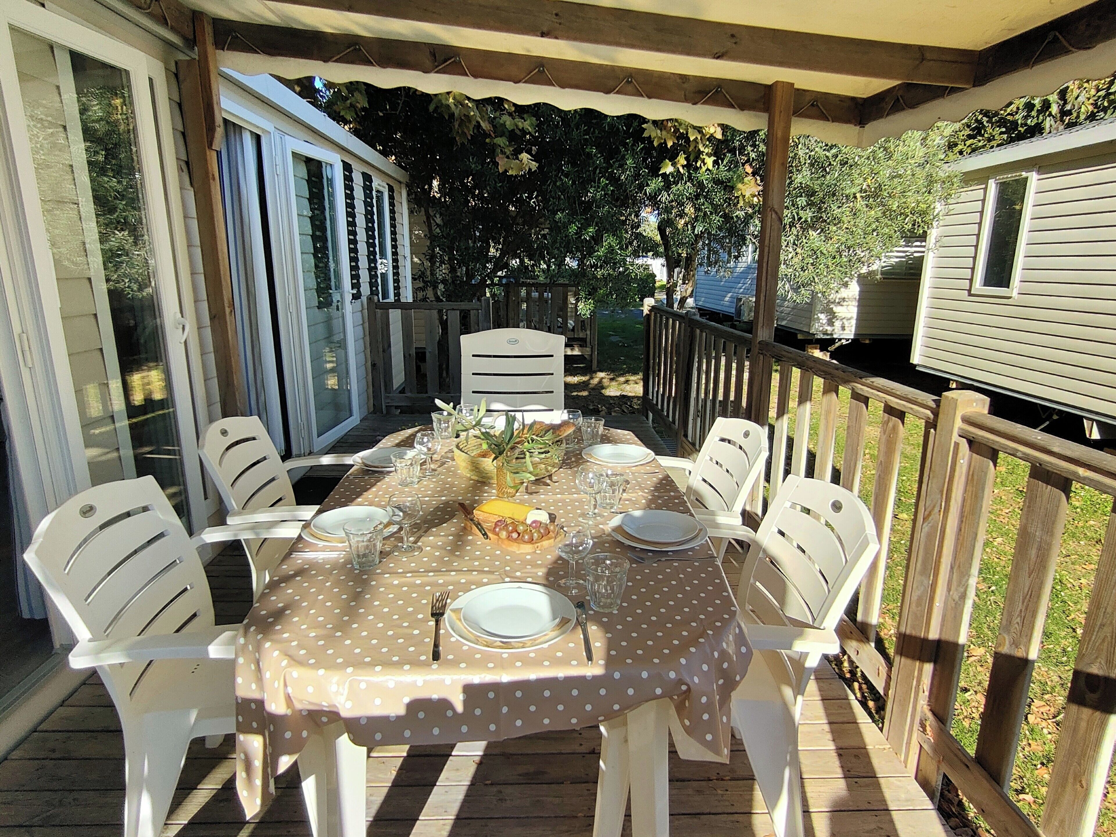 Un espace extérieur abrité, aménagé pour les repas, avec une table dressée pour quatre personnes, entourée de chaises blanches, et une terrasse en bois dotée d’une rambarde.