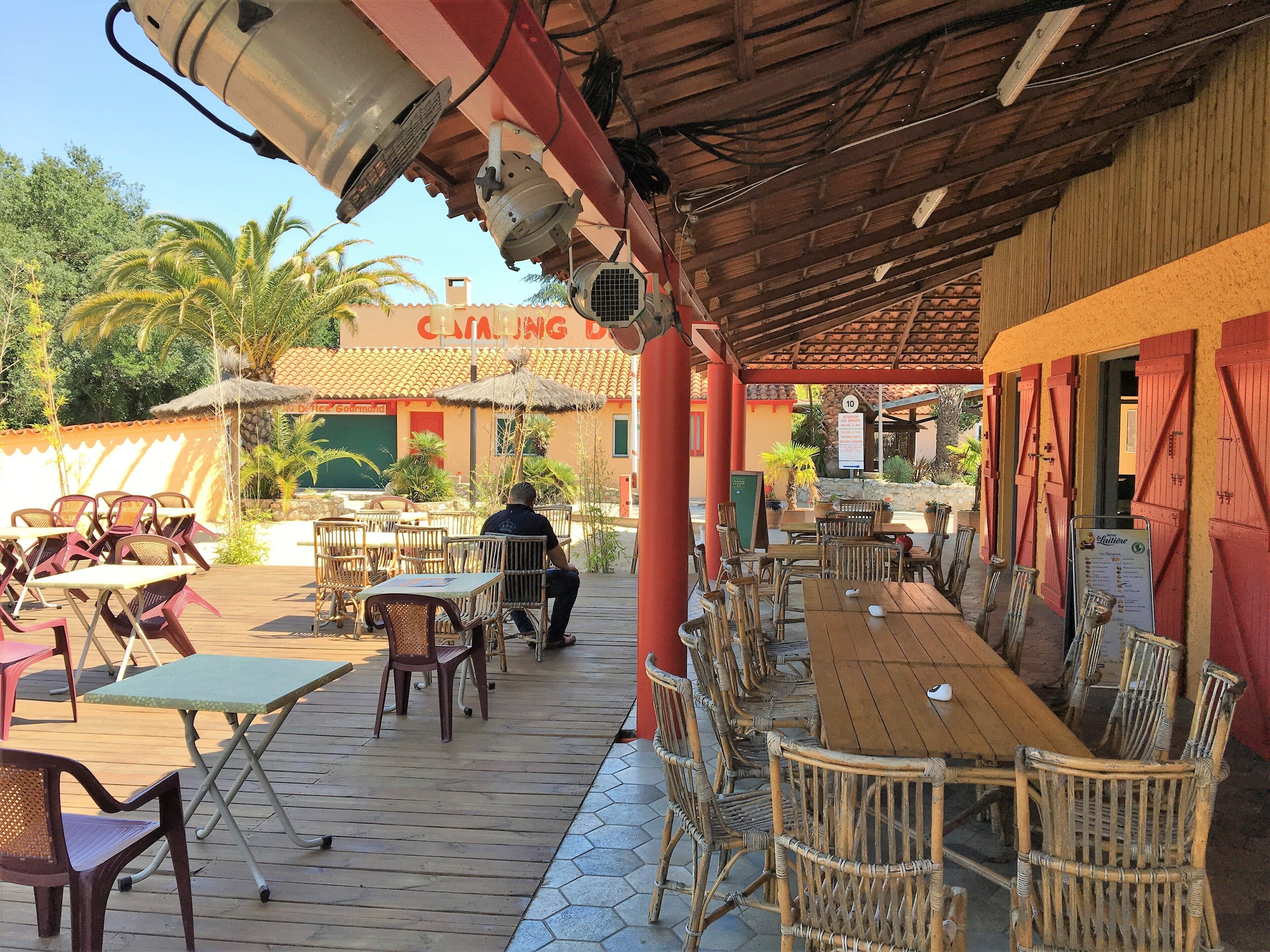 Un espace extérieur aménagé pour les repas, avec des tables et des chaises, une partie couverte et un bâtiment portant une enseigne.
