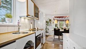 Een moderne keuken met witte kastjes, een houten aanrechtblad en een inbouwoven. Er is een raam met uitzicht op bomen en een eethoek met een tafel en stoelen.