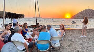 Een groep mensen zit aan een tafel aan het strand, met een ondergaande zon op de achtergrond.