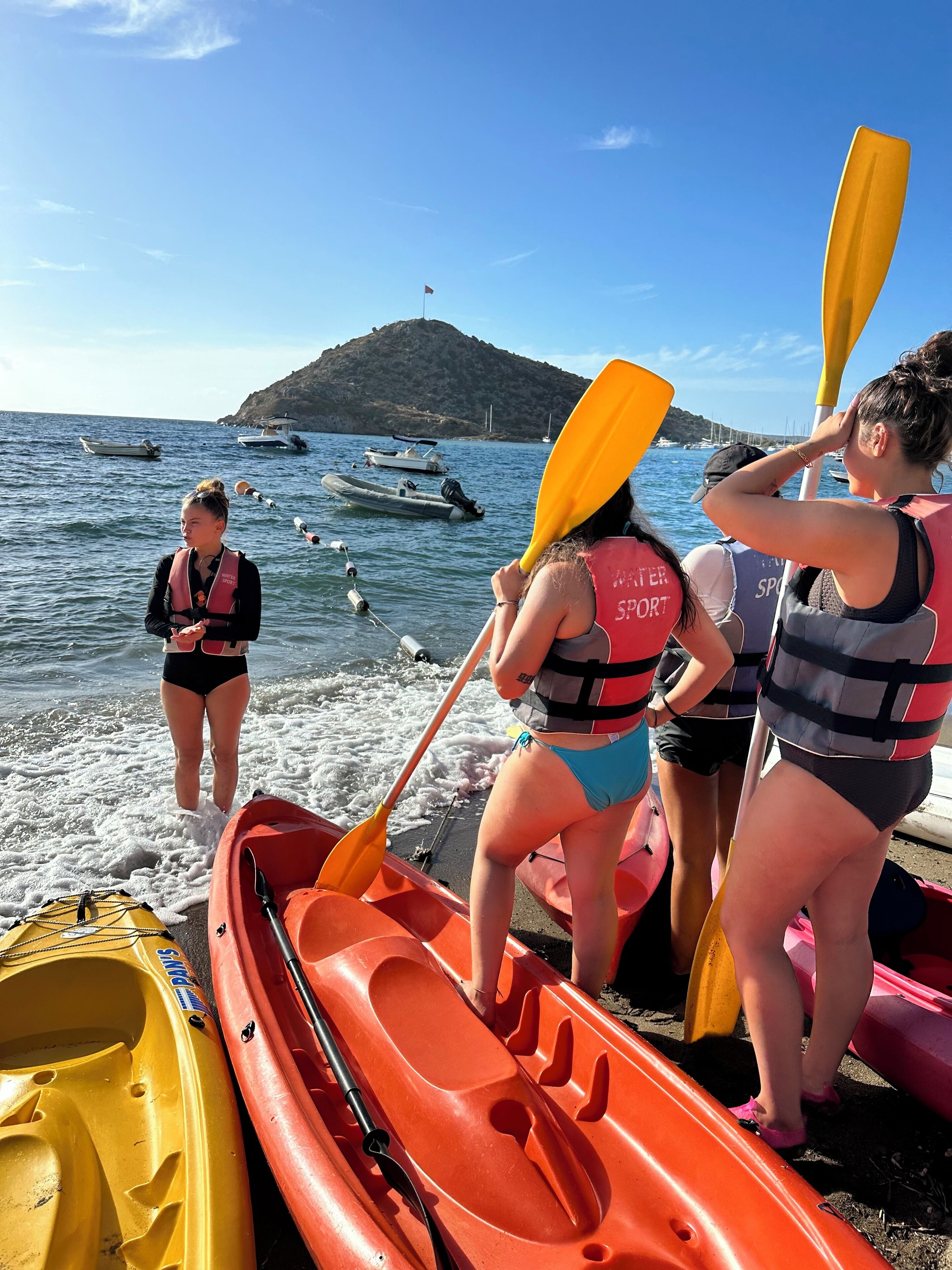 A group of people preparing kayaks on a beach with boats in the background.